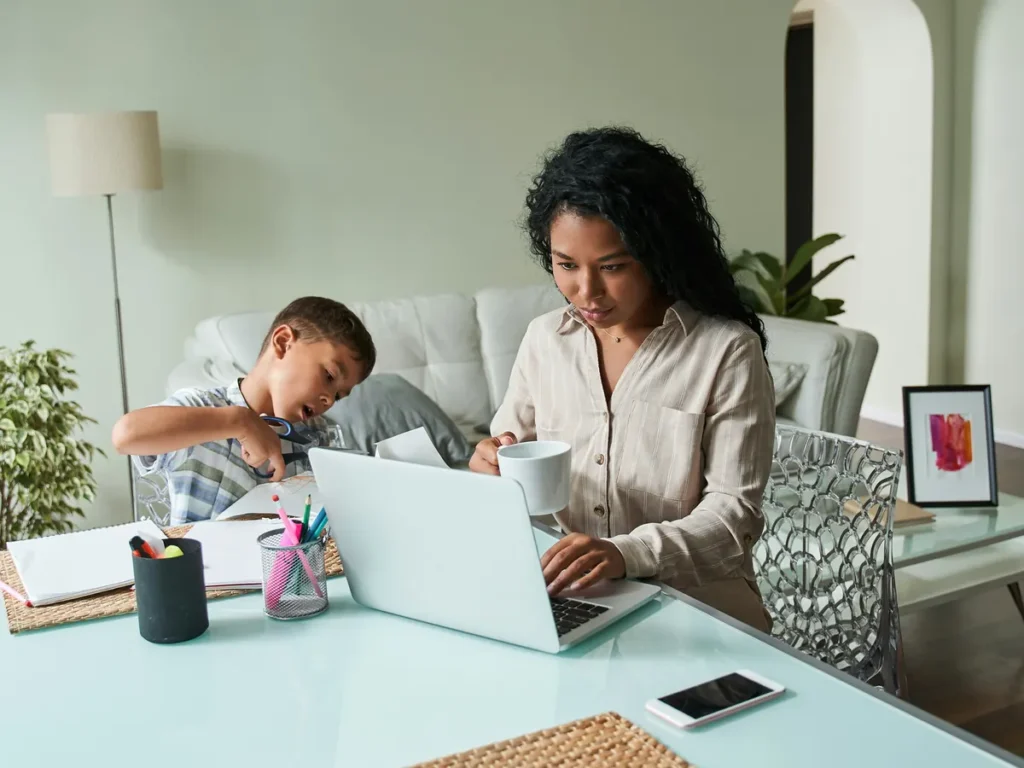 A mother working at her laptop while her child draws beside her at the table